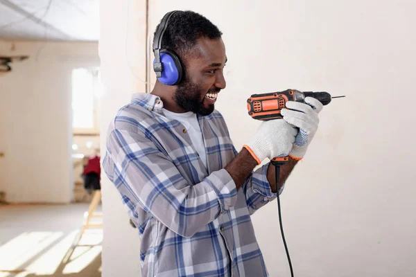 Side view of laughing young african american man in protective headphones and gloves having fun with power drill during renovation of home — Stock Photo