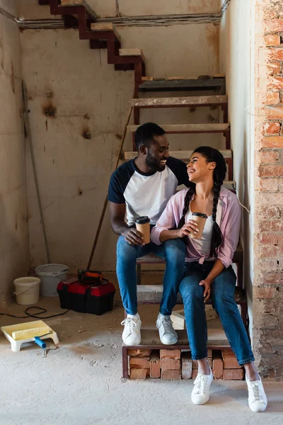 Sorridente coppia afro-americana a riposo con tazze di caffè usa e getta durante la ristrutturazione nella nuova casa — Foto stock