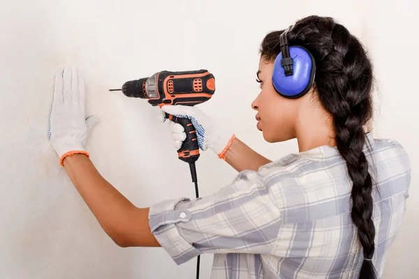 Femme afro-américaine concentrée dans des écouteurs de protection et des gants de travail avec perceuse électrique — Photo de stock