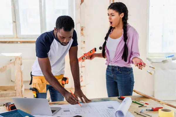 African american man making notes on blueprint at table with laptop while his angry girlfriend yelling at him near during renovation of home — Stock Photo