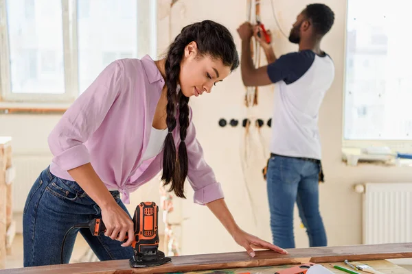 Selective focus of african american woman working with jigsaw while her boyfriend standing behind during renovation at home — Stock Photo