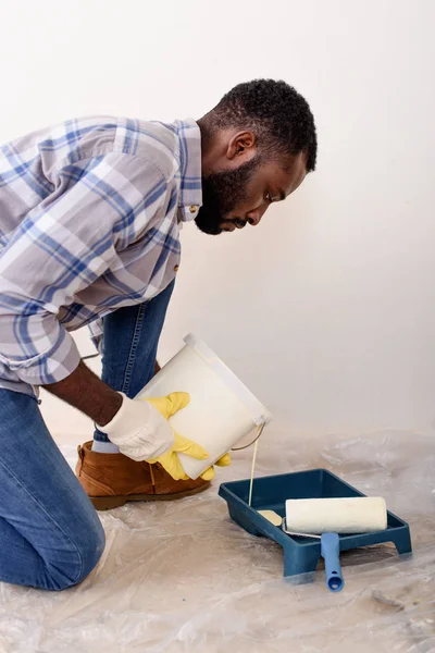 Hombre afroamericano enfocado en guantes protectores vertiendo pintura en bandeja de rodillos durante la renovación del hogar - foto de stock