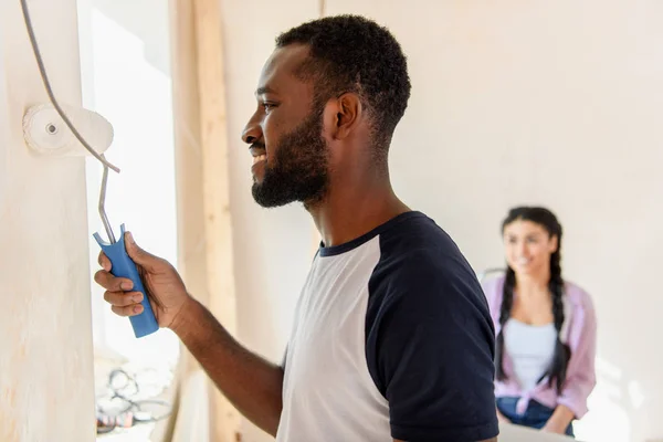 Vista lateral de hombre afroamericano feliz pintura pared por rodillo de pintura mientras que su novia sentada detrás en casa - foto de stock