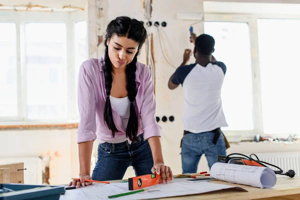 Pensive young woman with spirit level looking at blueprint while her boyfriend working behind during renovation of home — Stock Photo