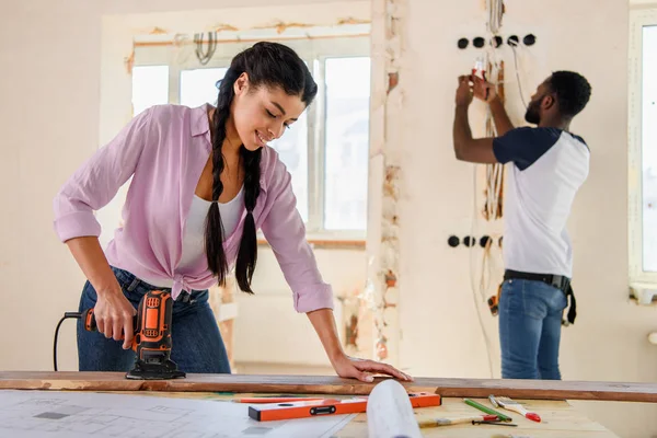 Attractive african american woman working with jigsaw while her boyfriend standing behind during renovation at home — Stock Photo