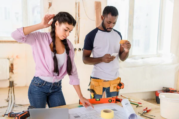 Young african american man with credit card and smartphone doing online shopping while his girlfriend looking at blueprint during renovation of home — Stock Photo
