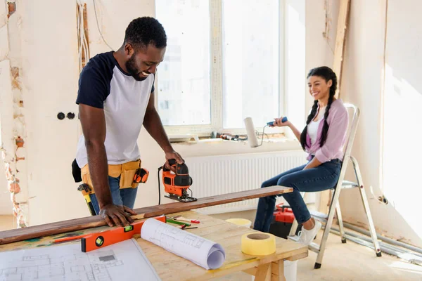 Sonriente hombre afroamericano trabajando con rompecabezas mientras su novia se sienta cerca durante la renovación en casa - foto de stock