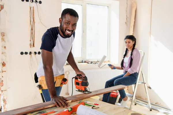 Handsome african american man working with jigsaw while his girlfriend sitting near during renovation at home — Stock Photo