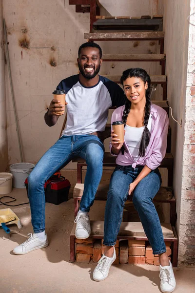 Feliz pareja afroamericana descansando con tazas de café desechables durante la renovación en un nuevo hogar - foto de stock