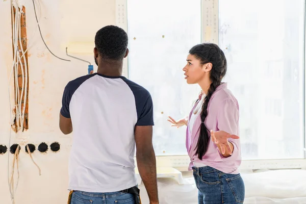 Vista trasera del hombre pintando pared por rodillo de pintura mientras su novia irritada haciendo gesto encogiéndose de hombros - foto de stock