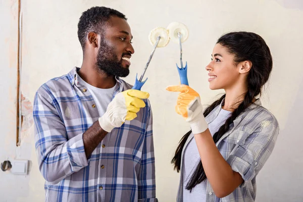 Sorrindo casal afro-americano segurando rolos de tinta e olhando uns para os outros durante a renovação em casa — Fotografia de Stock