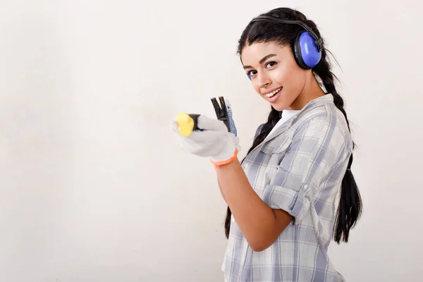 Feliz joven mujer en los auriculares de protección que se divierten con la herramienta y mirando a la cámara - foto de stock