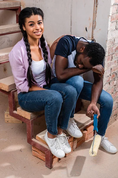 Високий кут зору привабливої афроамериканської жінки, що підбадьорює виснаженого хлопця з фарбою під час ремонту будинку — стокове фото