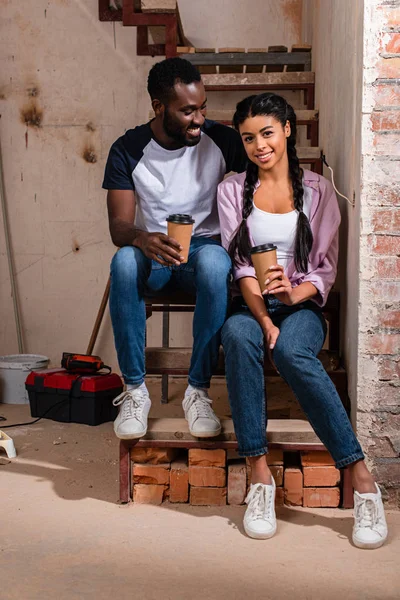 Alegre pareja afroamericana descansando con tazas de café desechables durante la renovación en el nuevo hogar - foto de stock