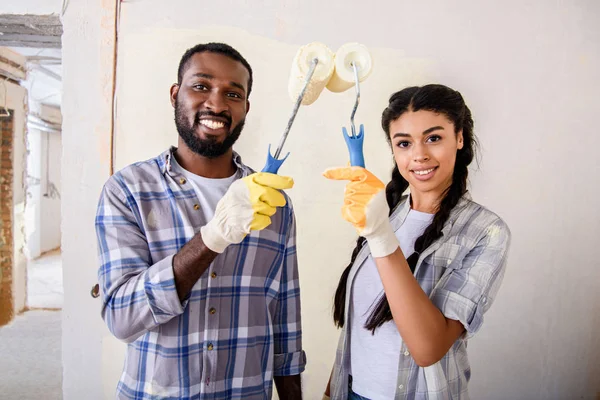 Alegre casal afro-americano segurando rolos de tinta e olhando para a câmera durante a renovação em casa — Fotografia de Stock