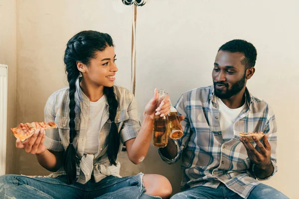 Feliz casal afro-americano com piza clinking por garrafas de cerveja durante a renovação em casa — Fotografia de Stock