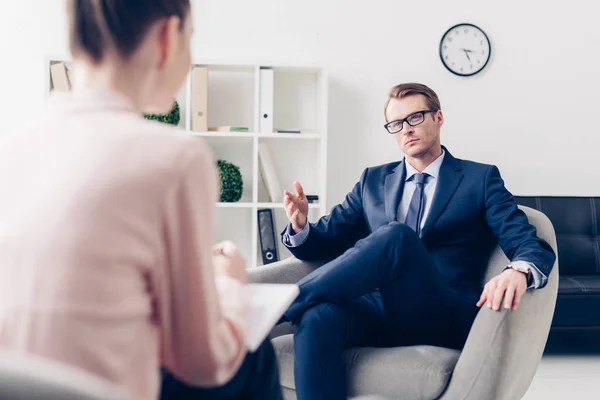 Enfoque selectivo de hombre de negocios guapo en traje y gafas dando entrevista a periodista en la oficina - foto de stock