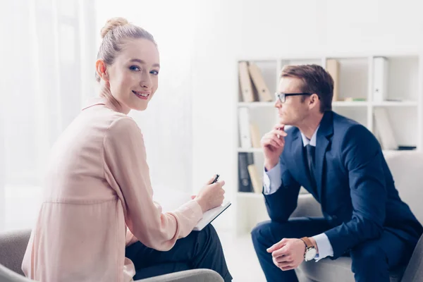 Fröhliche Journalistin macht sich Notizen während eines Interviews mit einem Geschäftsmann im Amt und blickt in die Kamera — Stockfoto