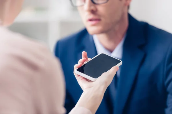 Imagen recortada de hombre de negocios dando entrevista a periodista con smartphone con pantalla en blanco en la oficina - foto de stock