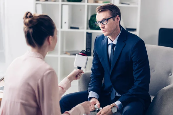 Handsome businessman in suit and glasses holding bottle of water and giving interview to journalist in office — Stock Photo