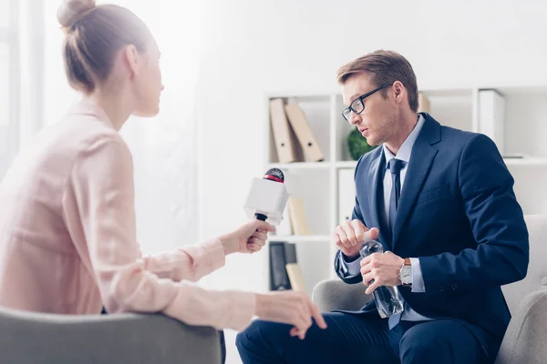 Bonito empresário segurando garrafa de água e dando entrevista para jornalista no escritório — Fotografia de Stock