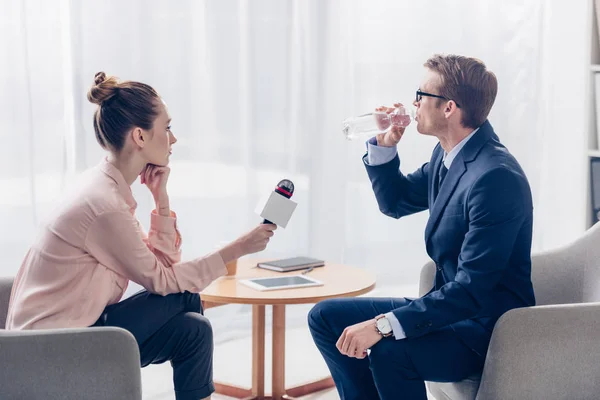 Vista lateral del hombre de negocios guapo beber agua mientras que da entrevista a periodista con micrófono en la oficina - foto de stock