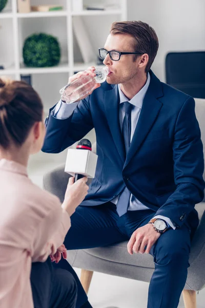 Apuesto empresario beber agua mientras dando entrevista a periodista con micrófono en la oficina - foto de stock