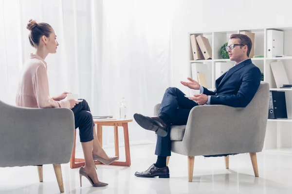 Vista lateral de hombre de negocios guapo dando entrevista a periodista en la oficina, hablando y sentado en sillones con tazas de café - foto de stock