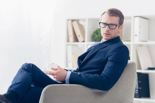 Seriöser Geschäftsmann in Anzug und Brille sitzt mit Tasse Tee im Sessel im Büro und blickt in die Kamera — Stockfoto