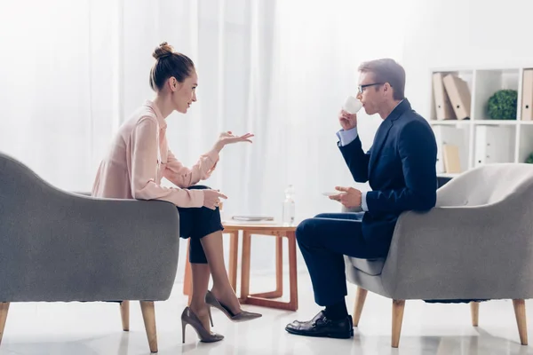 Seitenansicht eines attraktiven Journalisten, der Fragen stellt, während Geschäftsmann im Büro Kaffee trinkt — Stockfoto