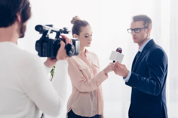 Bel homme d'affaires en lunettes donnant une interview vidéo au journaliste et cameraman au bureau — Photo de stock
