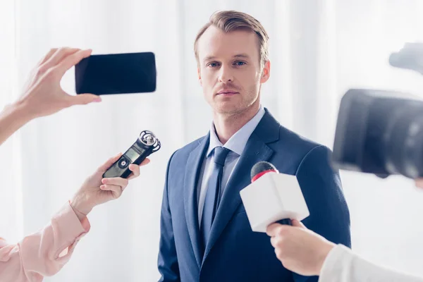 Apuesto hombre de negocios dando entrevista a periodistas y mirando a la cámara en la oficina - foto de stock