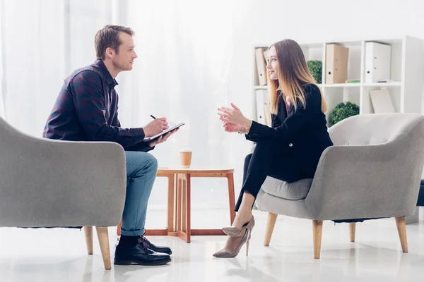 Vista laterale di allegra donna d'affari in giacca e cravatta seduta sulla poltrona e che rilascia intervista al giornalista in carica — Foto stock