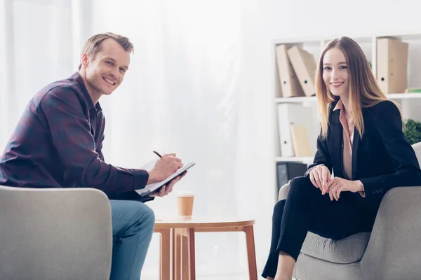 Smiling attractive businesswoman in suit and handsome journalist looking at camera in office — Stock Photo