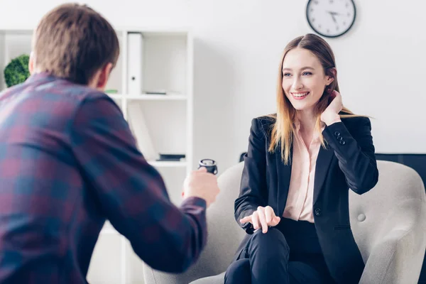 Smiling businesswoman in suit touching hair while giving interview to journalist in office — Stock Photo