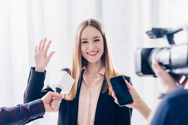 Heureuse femme d'affaires attrayante donnant une interview aux journalistes et agitant la main dans le bureau — Photo de stock