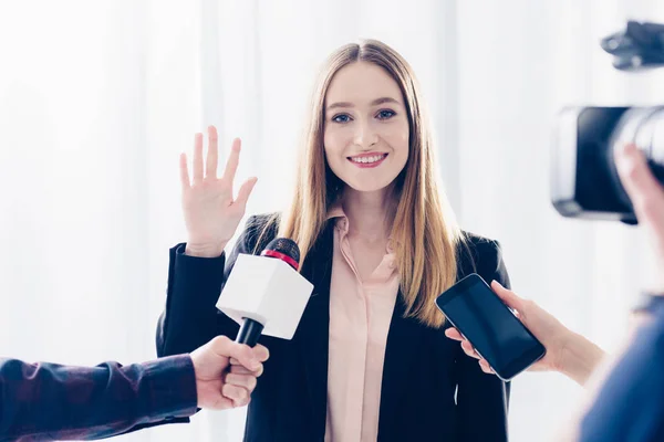 Sonriente atractiva empresaria dando entrevista a periodistas y saludando de la mano en el cargo - foto de stock