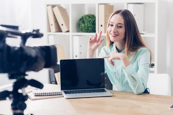 Smiling attractive female video blogger recording vlog in office, laptop with blank screen on tabletop — Stock Photo