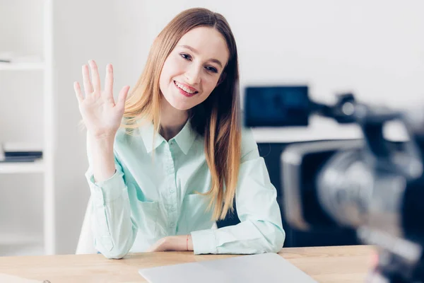 Усміхнений привабливий жіночий відео блогер записує відеоблог і махає рукою в офісі — стокове фото