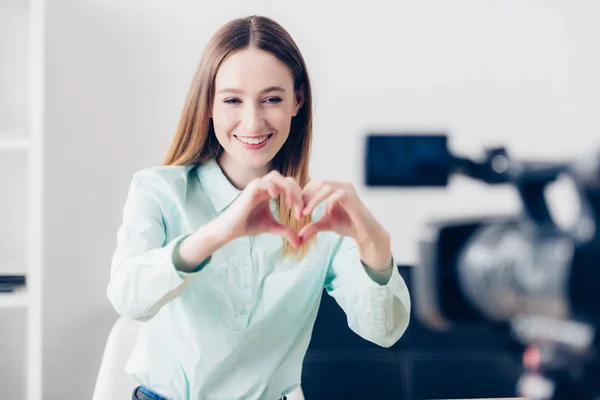 Happy attractive female video blogger recording vlog and showing heart with fingers in office — Stock Photo