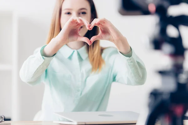 Selective focus of attractive female video blogger recording vlog and showing heart with fingers in office — Stock Photo