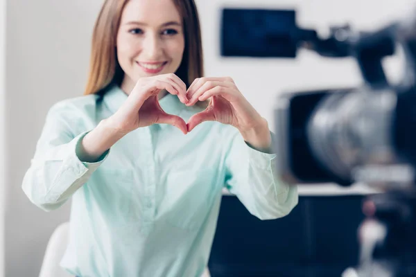 Smiling attractive female video blogger recording vlog and showing heart with fingers in office — Stock Photo