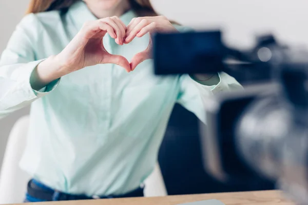 Cropped image of female video blogger recording vlog and showing heart with fingers in office — Stock Photo