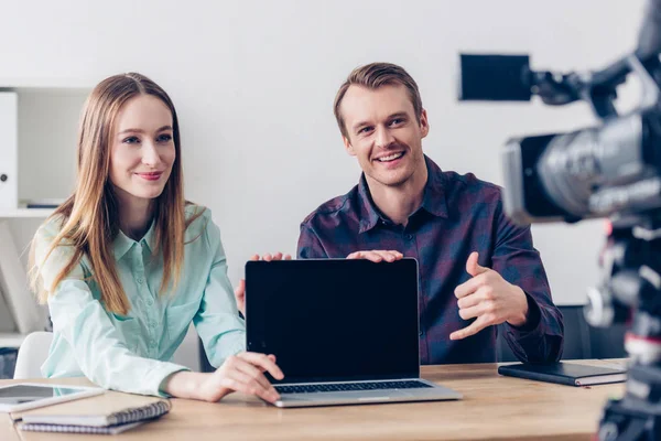 Happy video bloggers recording vlog and pointing on laptop with blank screen in office — Stock Photo