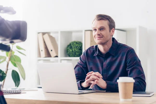 Усміхнений красивий чоловічий відео блогер записує відеоблог і сидить за столом з ноутбуком в офісі — стокове фото