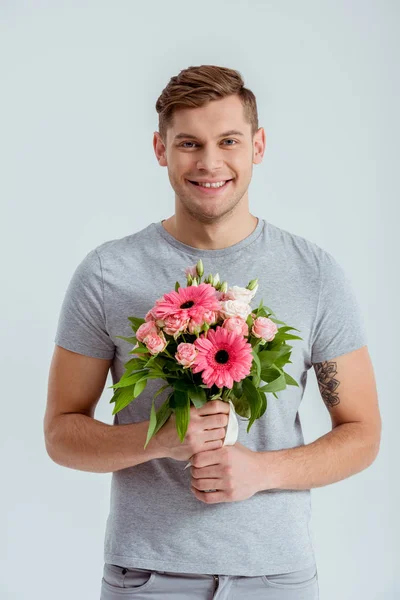 Hombre sonriente mirando a la cámara y sosteniendo ramo de flores rosa aislado en gris - foto de stock