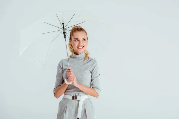 Усміхнена жінка в сірому одязі позує з прозорою парасолькою ізольовано на сірому — стокове фото