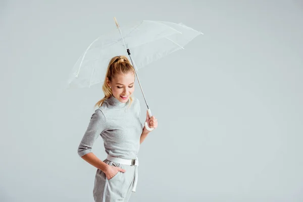 Femme souriante en vêtements gris et la main dans la poche posant avec parapluie transparent isolé sur gris — Photo de stock