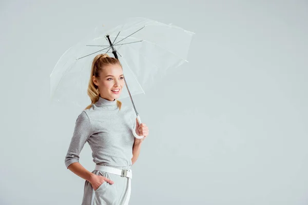 Hermosa mujer en ropa gris y mano en bolsillo posando con paraguas transparente aislado en gris - foto de stock