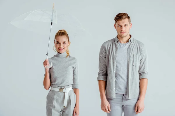 Belle femme souriante tenant parapluie transparent et debout près de l'homme insatisfait isolé sur gris — Photo de stock